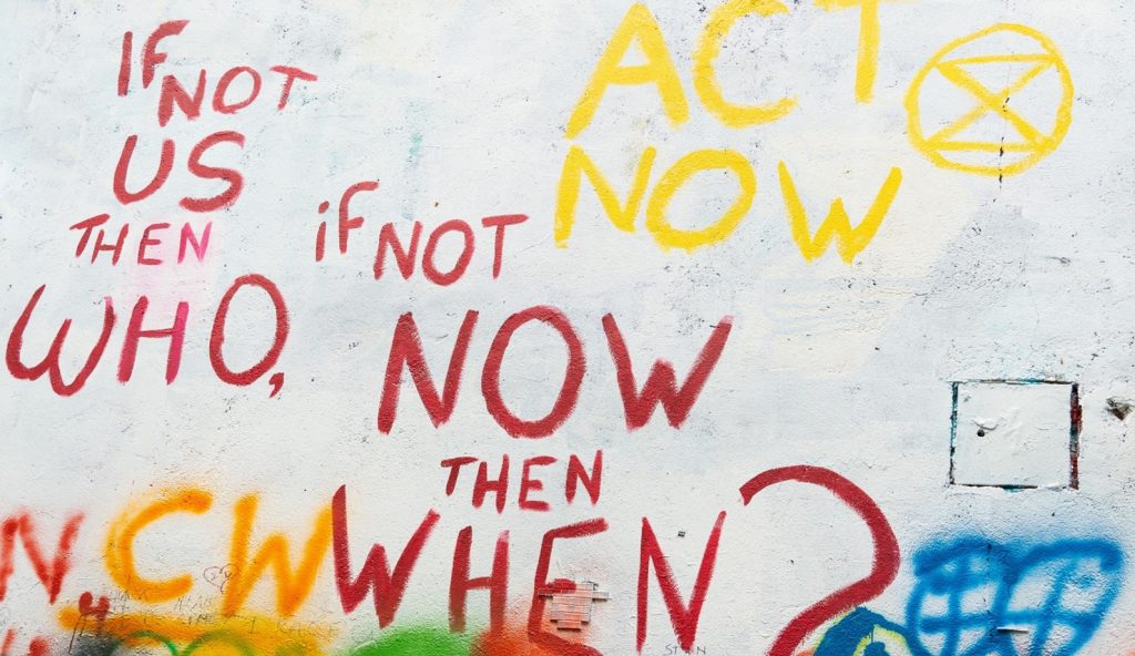 : Words in coloured graffiti on a wall that say “If not us then who, if not now then when? Act Now"