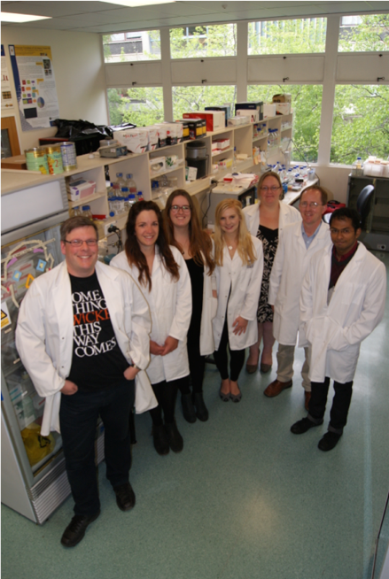 Photo of Professor Peter Dearden standing with six young researchers in a laboratory, all dressed in white lab coats.