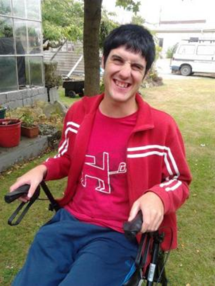 A smiling young man in a red top sits in a wheelchair in a backyard.