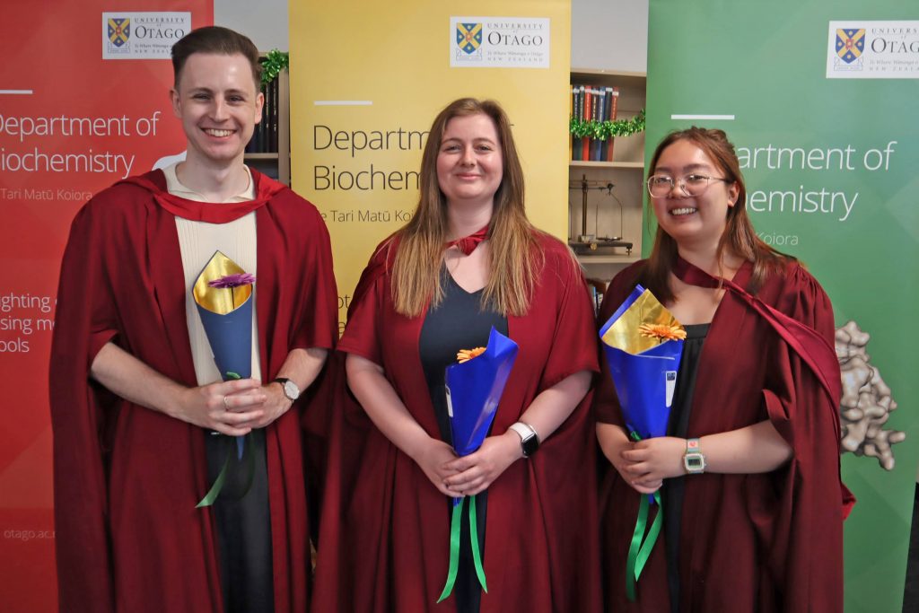 Three young adults wearing scarlet robes and holding gerbera flowers stand in front of red, yellow and green banners.