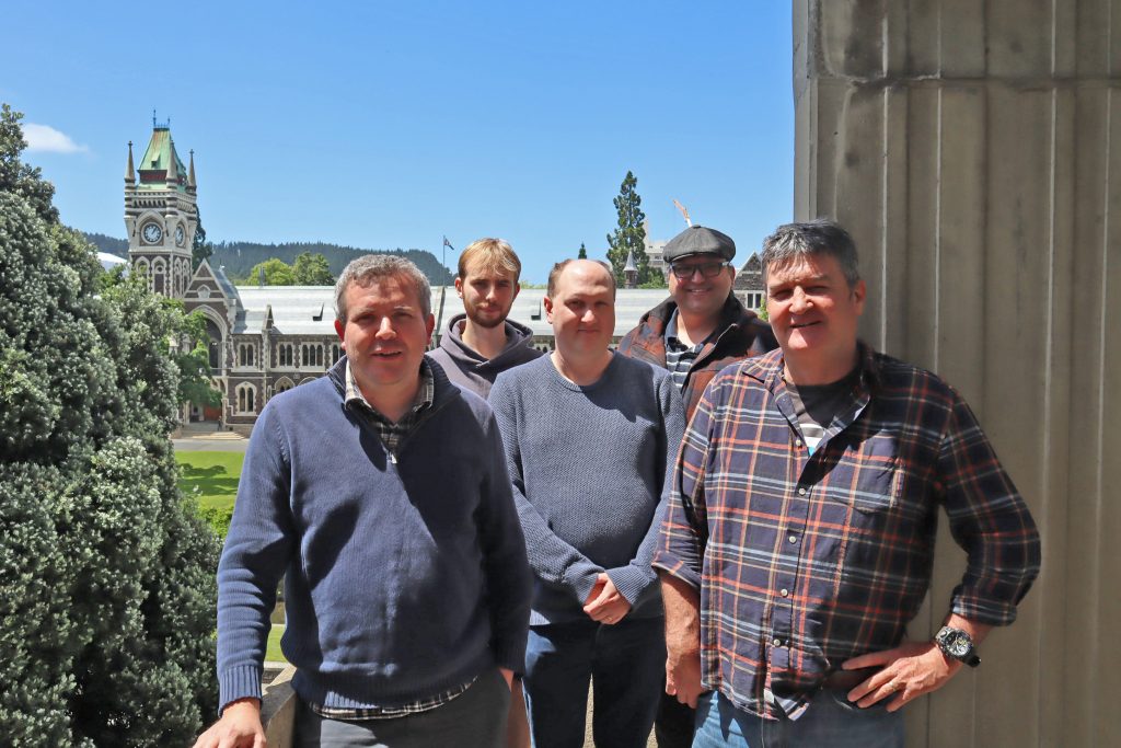 Five men stand in the sunshine with a tree to the left, a concrete wall on the right, and an elaborate clocktower building in the background.